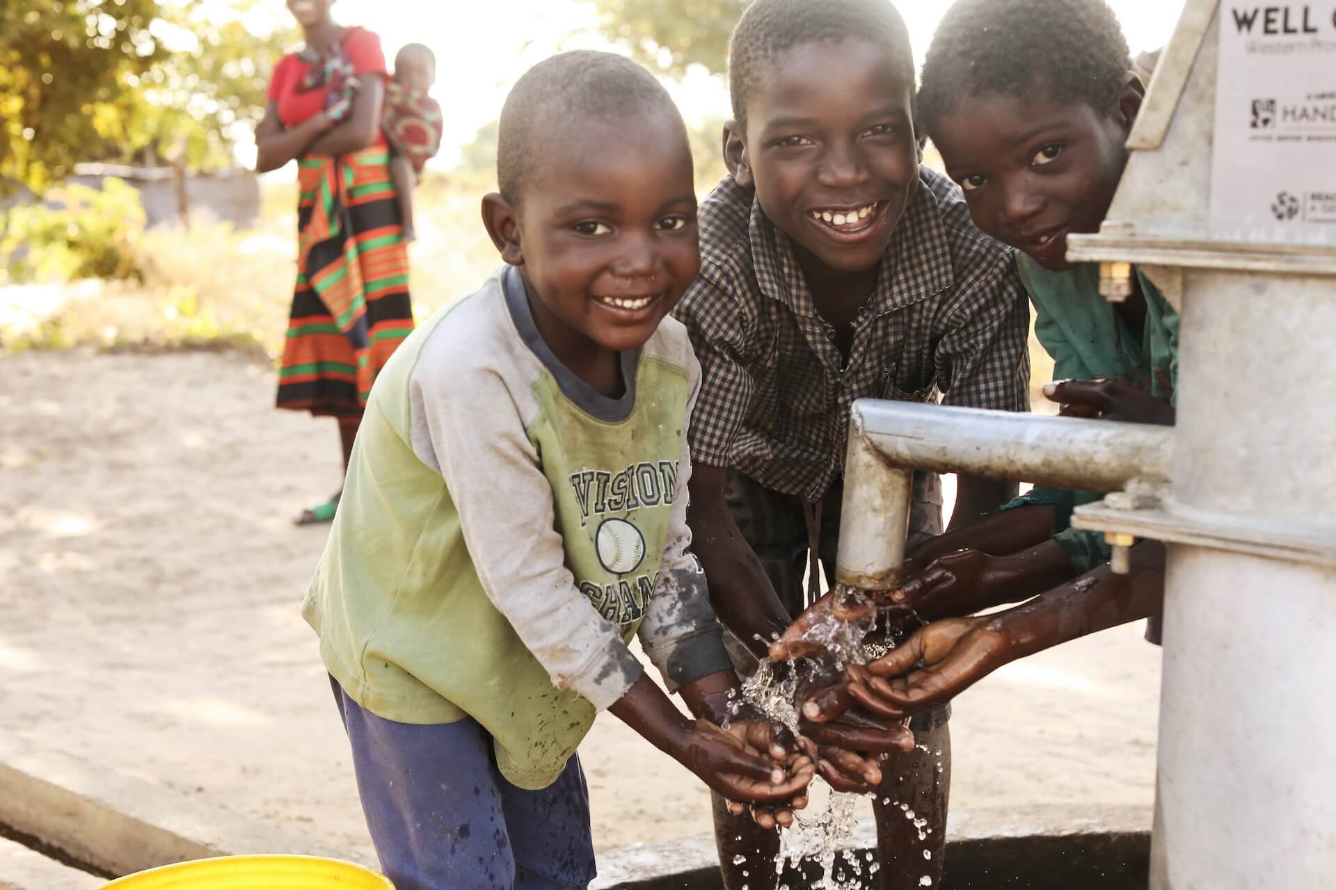 236A2917-6 (1) (1) Three smiling children wash their hands at a pump, collecting clean water. A woman holding a baby stands in the background, watching. The scene is outdoors in bright sunlight.