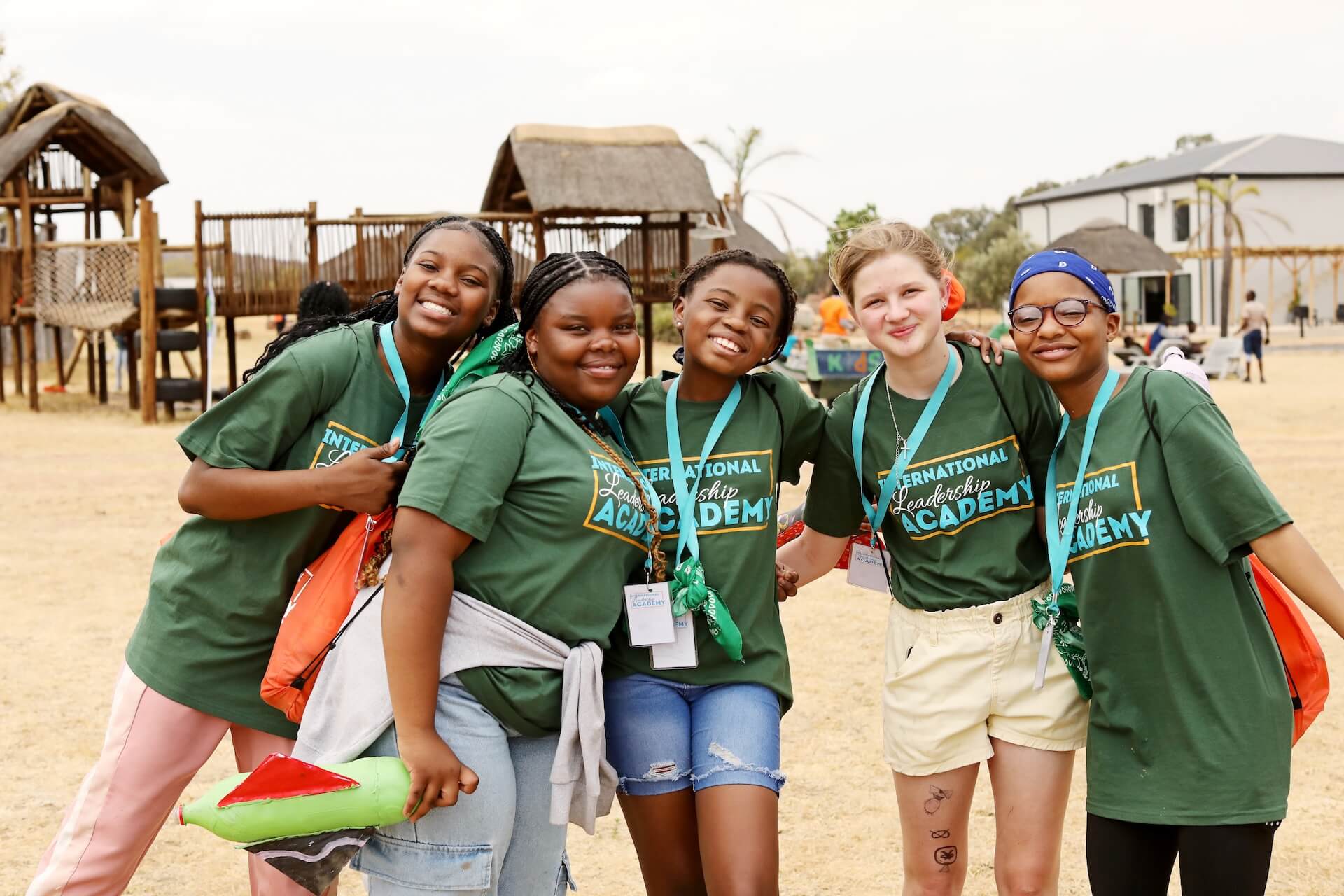 236A5724 (1) Five smiling young people in green International Leadership Academy shirts pose together outdoors, wearing name tags and lanyards, with playground equipment and huts visible in the background.