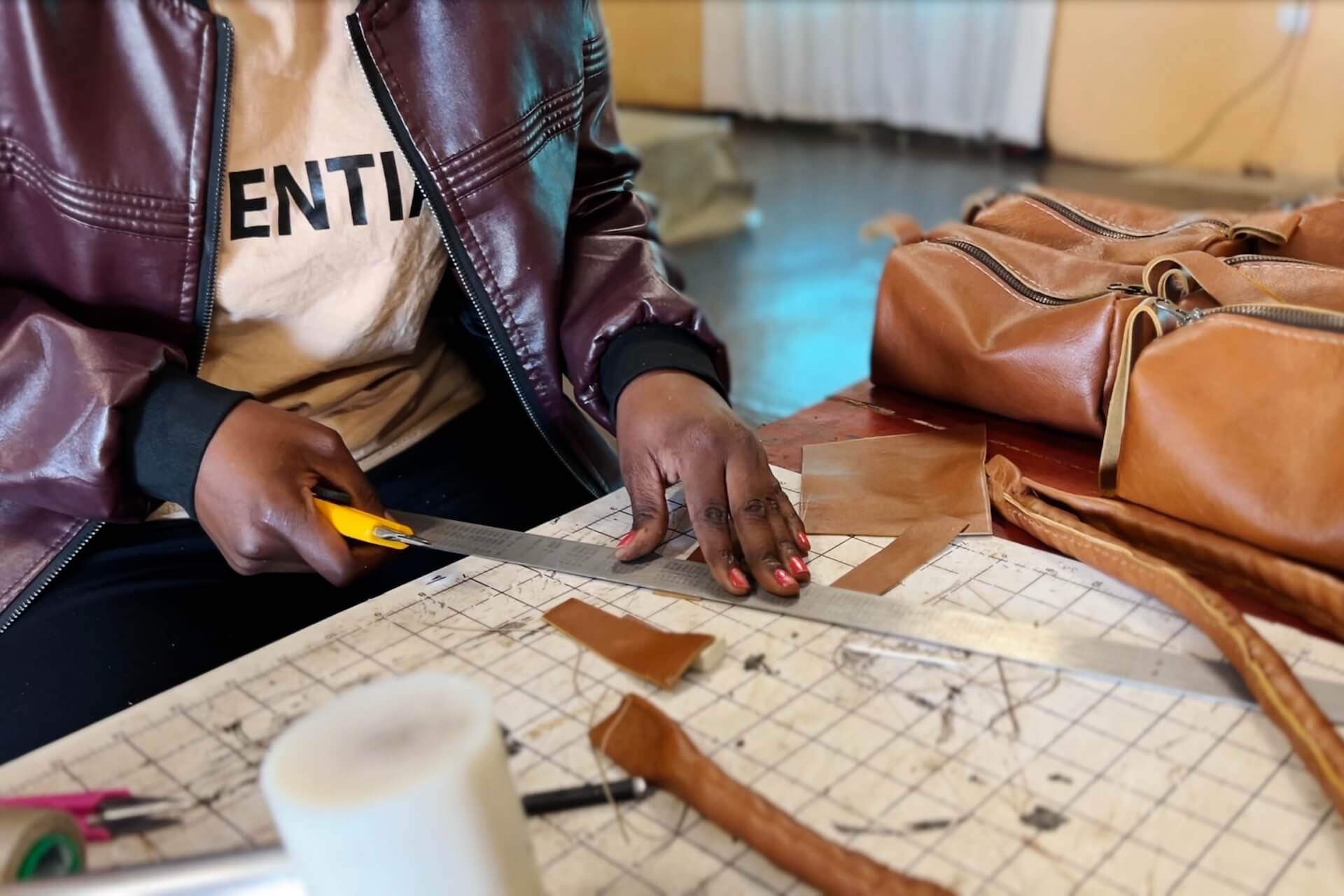 3 (1) A person in a maroon jacket cuts brown leather with a yellow utility knife and a metal ruler on a gridded workspace, with leather bags and scraps nearby.