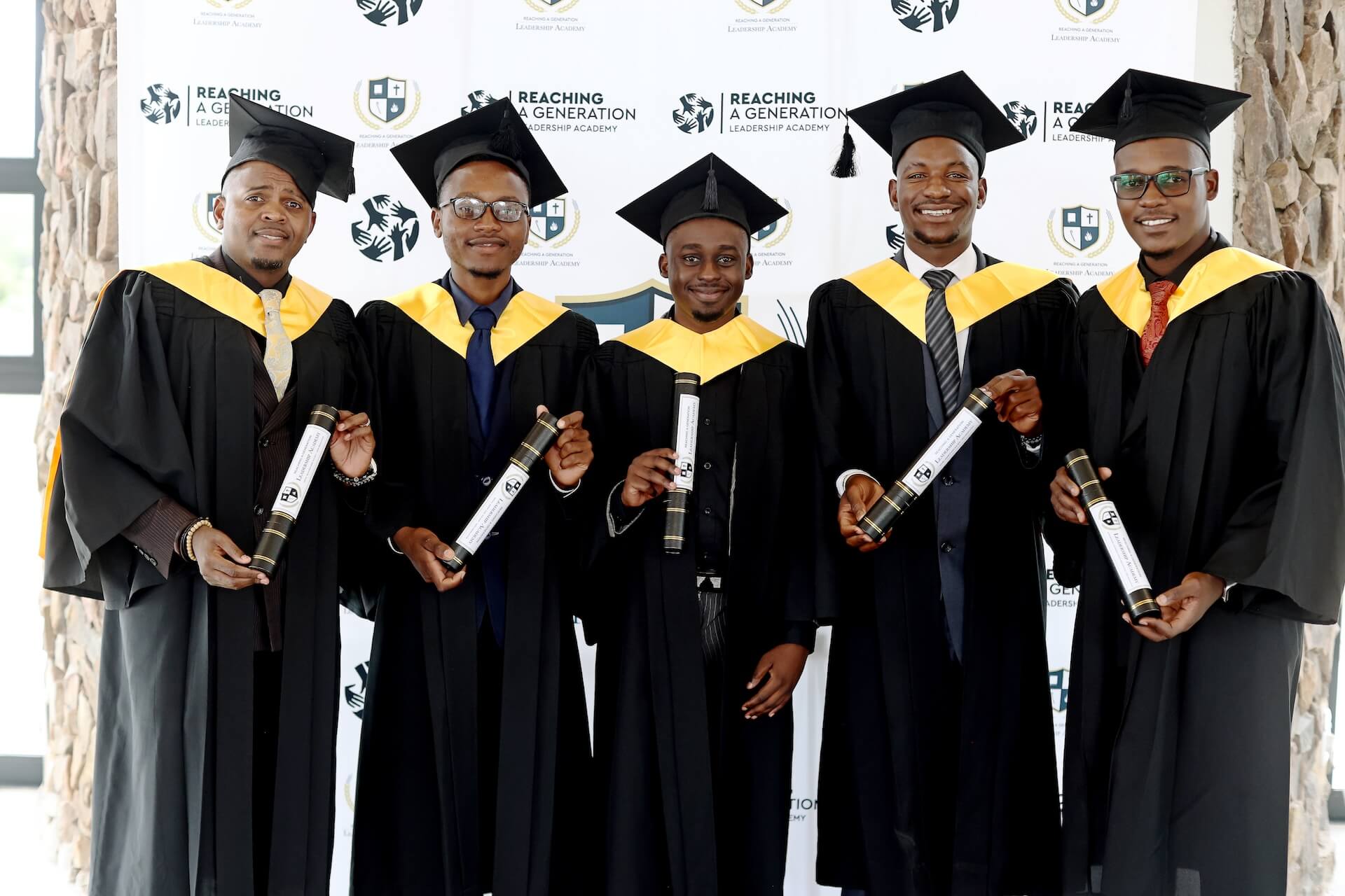 IMG_5110 Five men in graduation gowns and caps stand smiling, each holding a diploma, in front of a backdrop with Reaching a Generation logos.