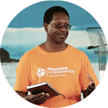 A man in glasses and an orange Reaching A Generation shirt holds a notebook while speaking, with a blue background behind him.