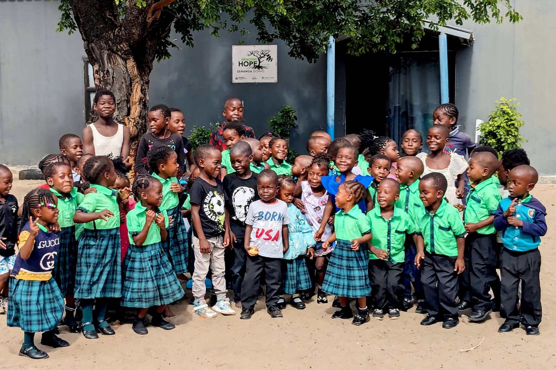 WhatsApp Image 2026-03-18 at 10.04.55-2 (1) A group of young children, some in green school uniforms and others in casual clothes, stand outside near a tree, smiling and gathered together with a few adults. A sign reading HOPE is visible on the building behind them.
