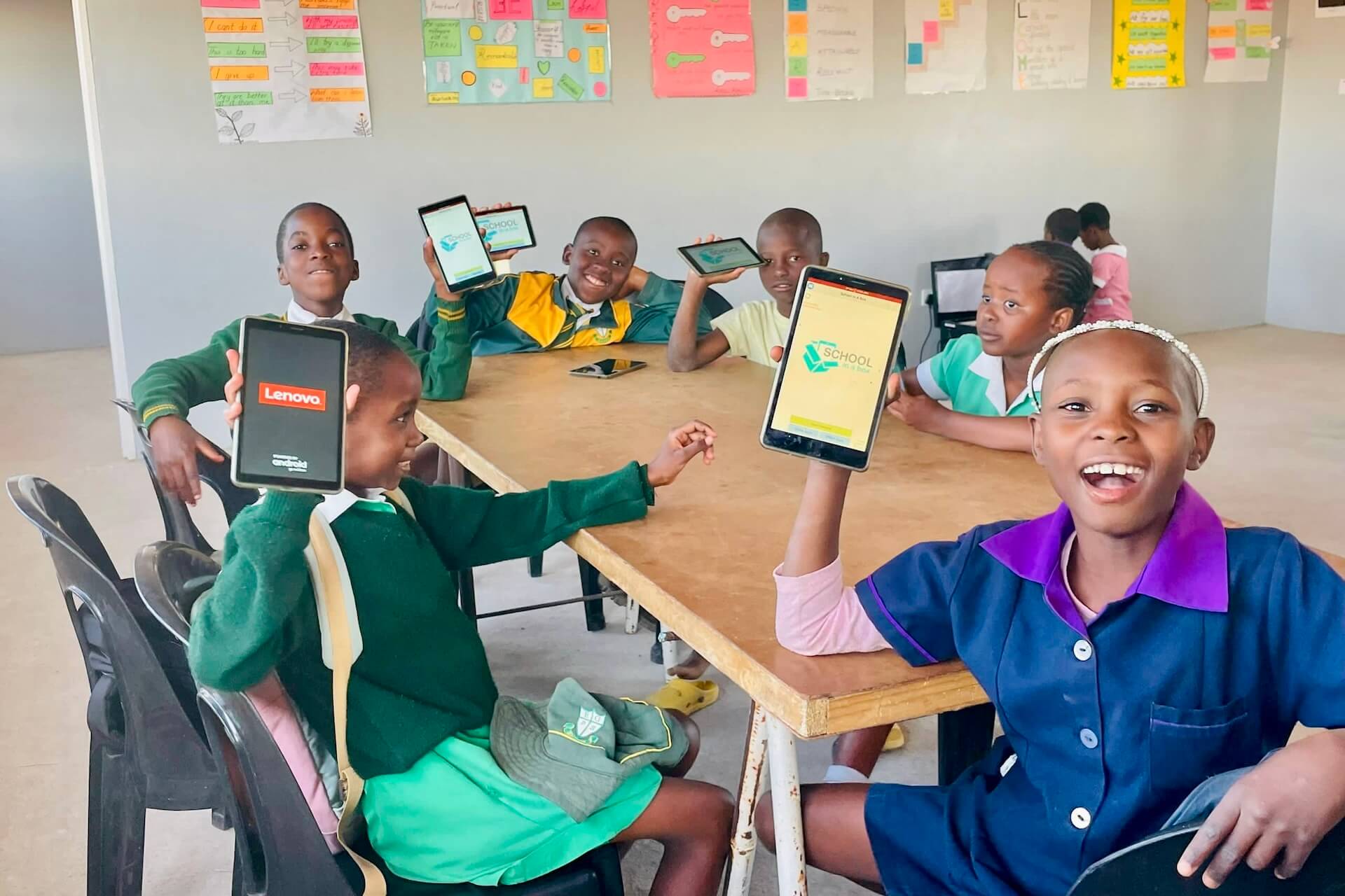 dda044c2-5c2d-4013-9986-8265d9ff6dc1 (2) Six children sit around a table in a classroom, smiling and holding up tablets. Colorful posters are on the wall behind them. The children wear school uniforms in green and blue.