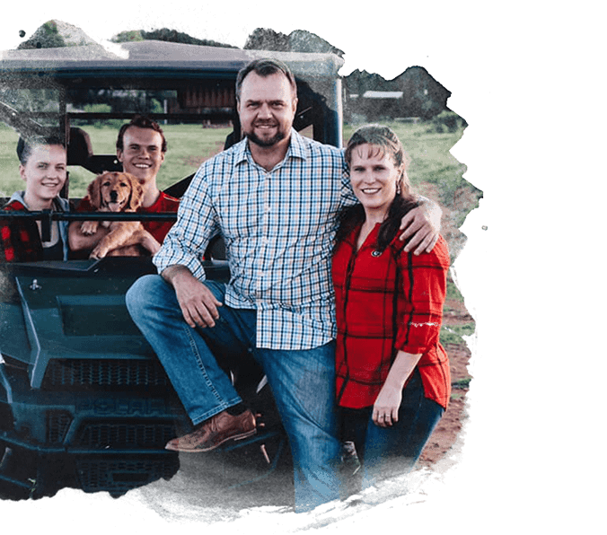 section-3 copy (1) Four people and a dog pose together outdoors by a green vehicle. Two people stand in front, while two and the dog sit on the back. All are smiling, and the scene has a painted or brushed-edge effect.
