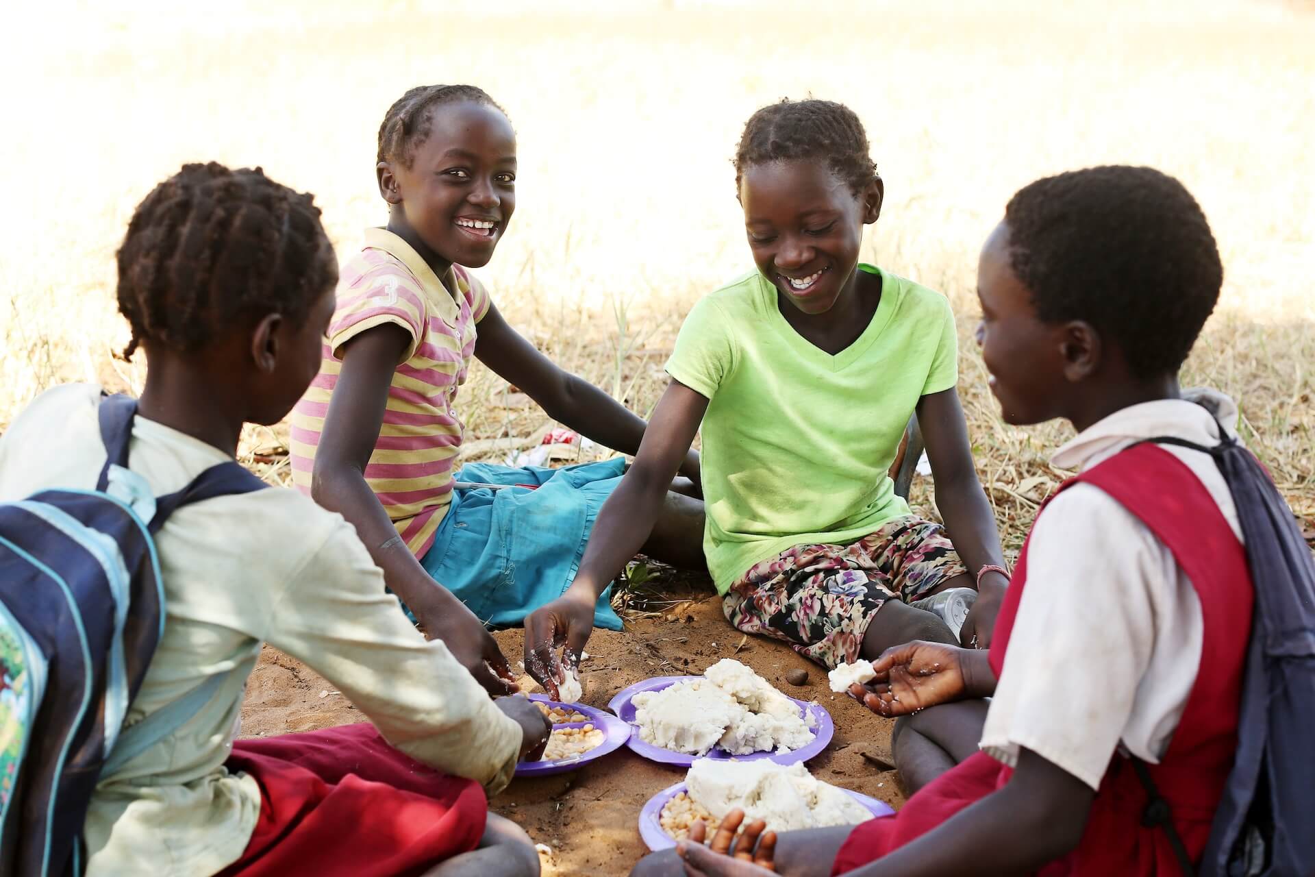 4.20.17_Zambia_month_trip_033 Four children sit on the ground outdoors, sharing food from plates and smiling together. They appear to be enjoying each others company in a natural, sunny setting.