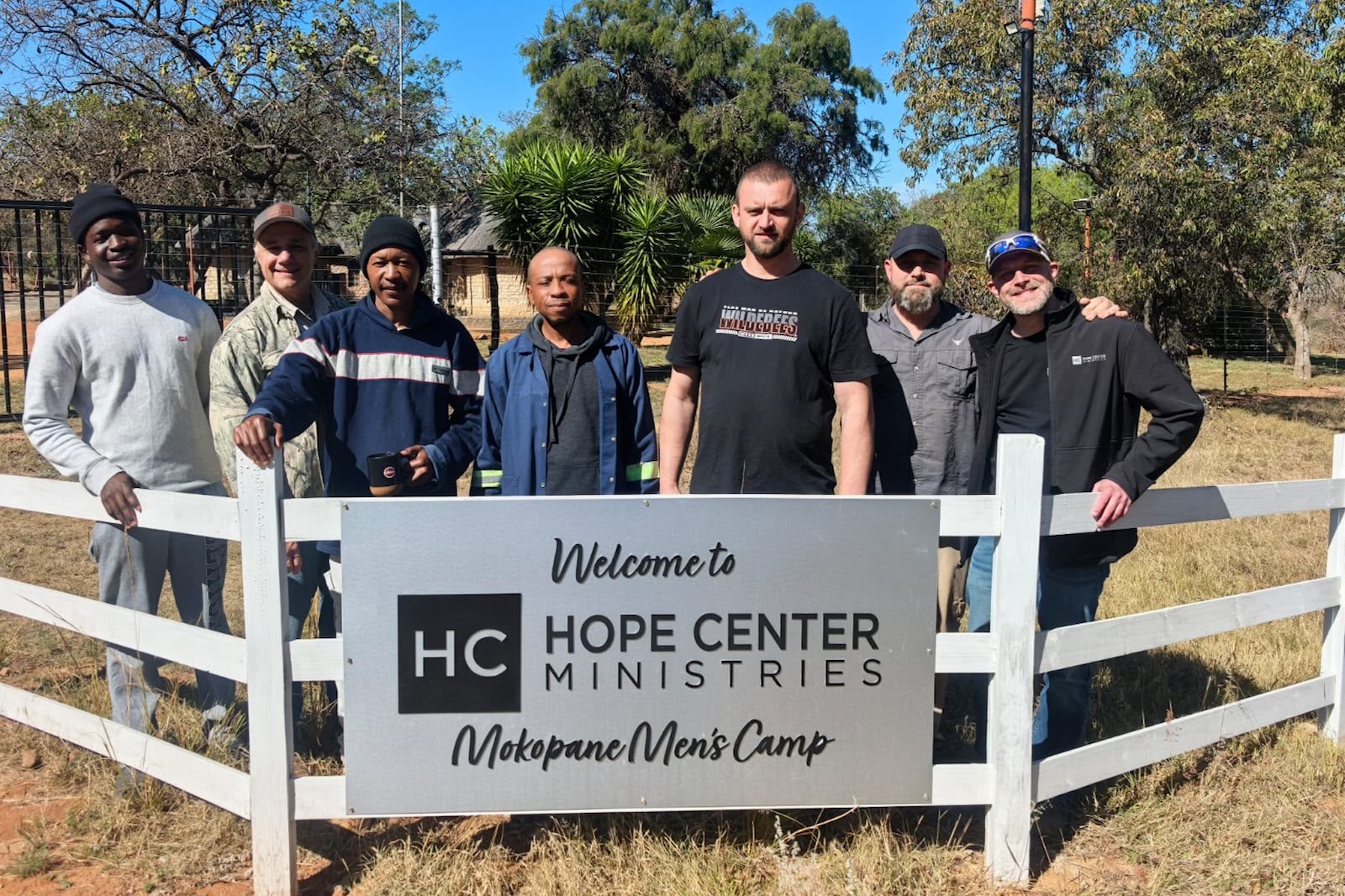 90ed699e-17f1-4806-9919-fcd5974b6b27 Seven men stand behind a white fence with a sign that reads Welcome to Hope Center Ministries Mokopane Mens Camp, with greenery and trees in the background.