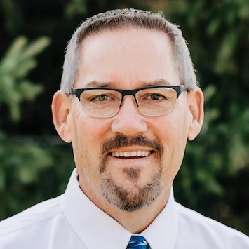MG_4966-e1532530331634-1 copy 2 Smiling man with short brown hair, glasses, and a goatee, wearing a white shirt and striped blue tie, standing in front of green foliage.